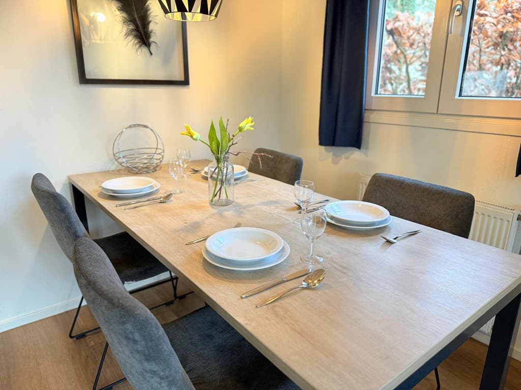 Eettafel met vier borden, glazen en bestek, bij een raam met uitzicht op de natuur.