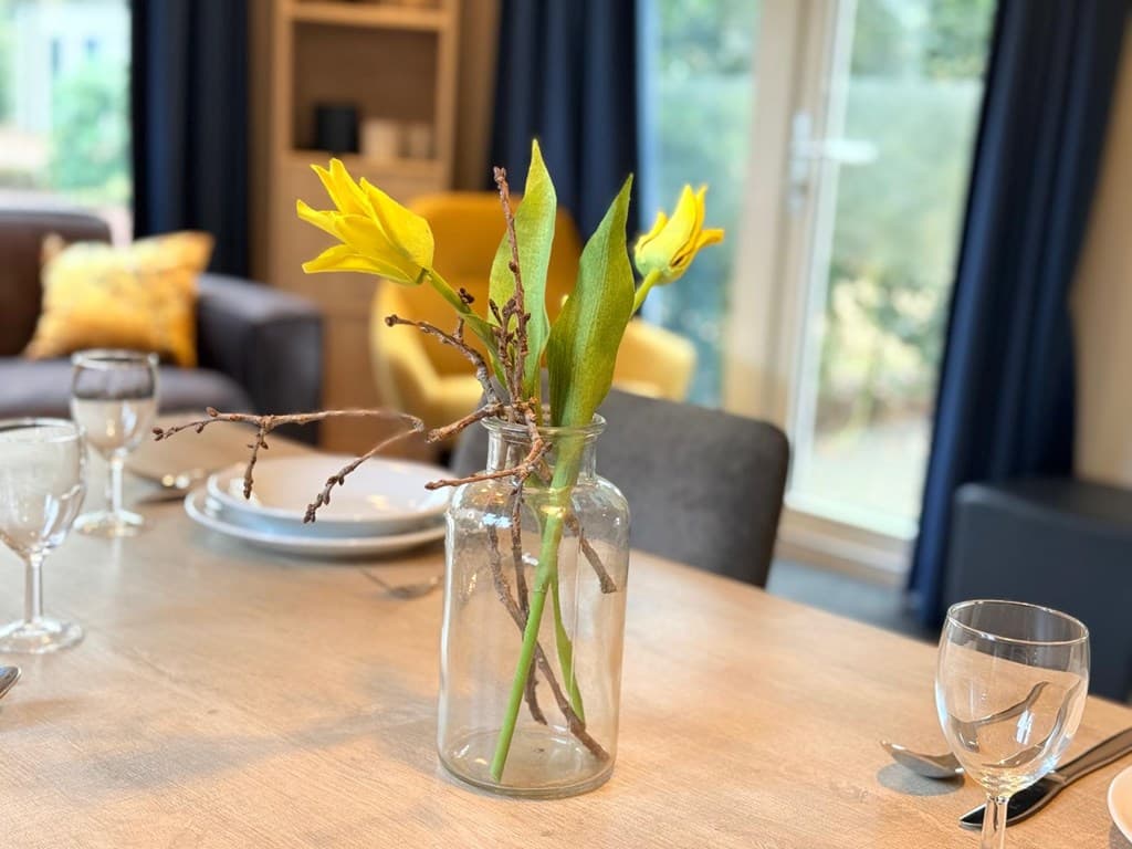 Kamer met een houten eettafel, glazen servies en een vaas met gele bloemen op de tafel.
