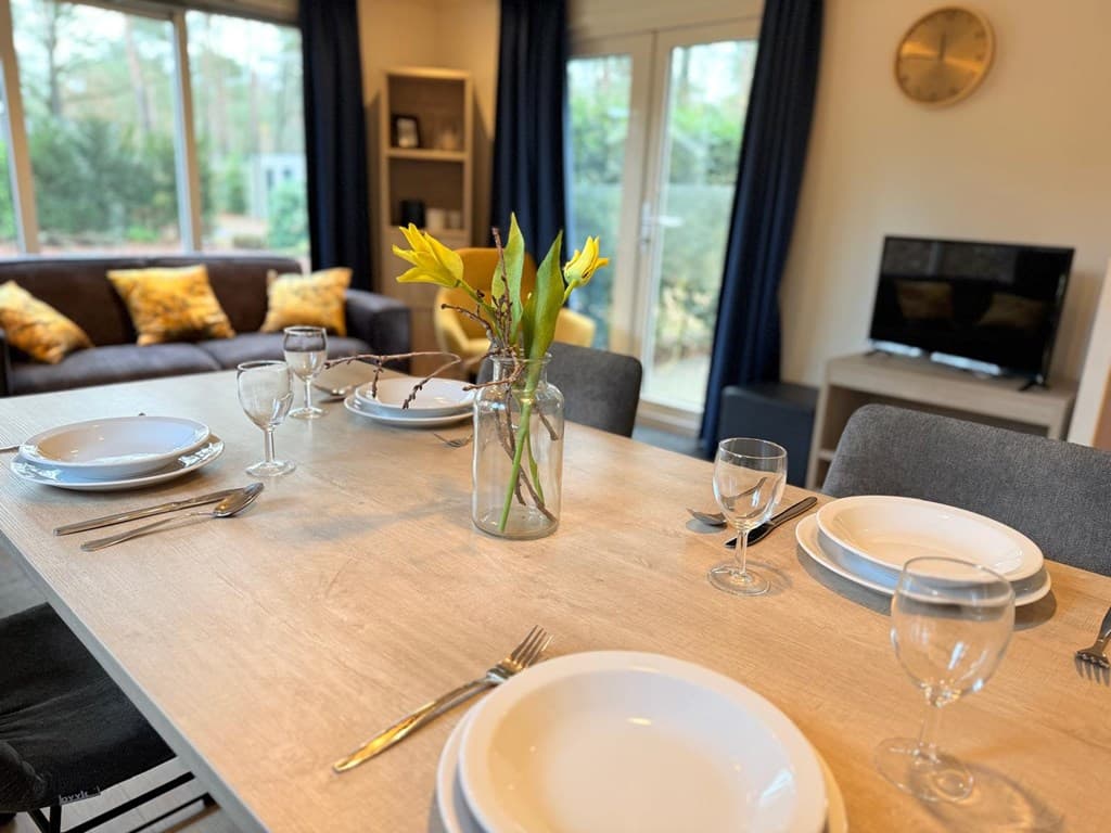 Eettafel met opgemaakte borden, glazen en een vaasje met bloemen, in een lichte woonkamer met zithoek en tv meubel