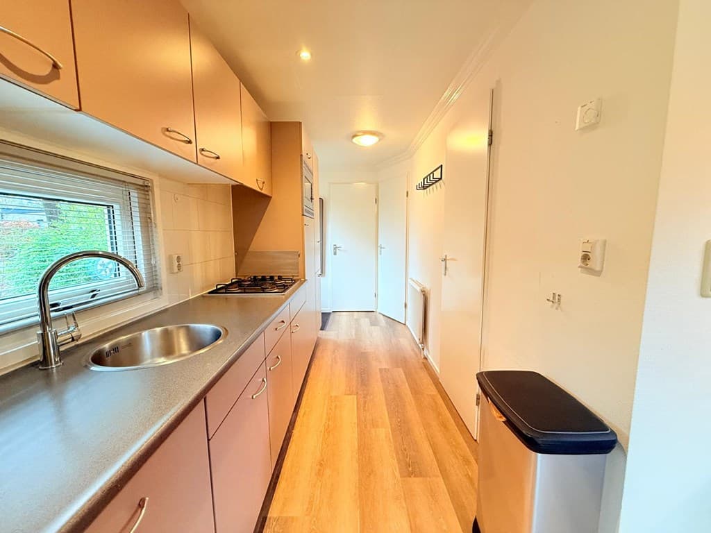 Keuken met inbouwkasten, een spoelbak, fornuis en toegang tot andere ruimtes. Houten vloer en lichtinval vanuit het raam.