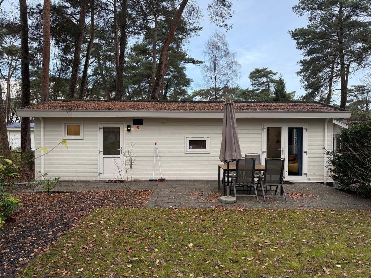 Zijaanzicht van een vakantiewoning met een terras, tafel en stoelen, omgeven door bomen en groen.