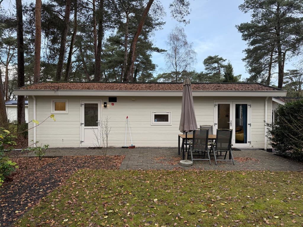 Zijaanzicht van een vakantiewoning met een terras, tafel en stoelen, omgeven door bomen en groen.