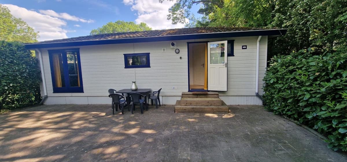 Witte vakantiewoning met een voordeur en terras met tafel en stoelen, omgeven door groen.