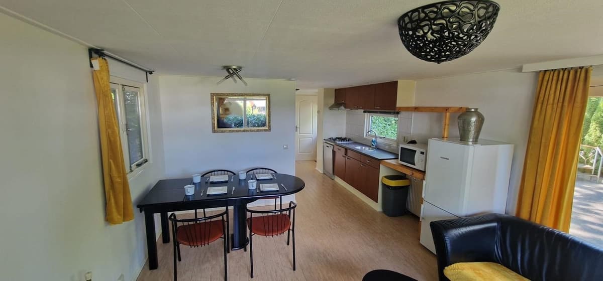 Beige woonkamer met een eettafel, keuken met apparatuur en een zithoek met een bank en grote ramen naar buiten