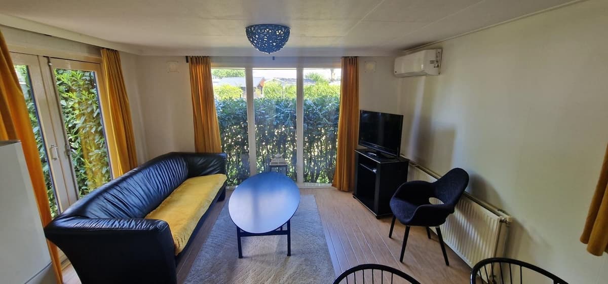 Woonkamer met een zwarte bank, eettafel en tv, grote ramen met zicht op een groene haag en gele gordijnen