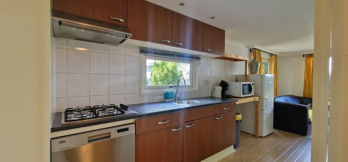 Moderne keuken met gasfornuis, spoelbak en inbouwapparatuur, naast een zitgedeelte met een bank en ramen met uitzicht.