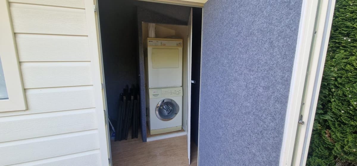 Ingang naar een berging met een wasmachine en droger, grijze wanden en opbergmaterialen zichtbaar.