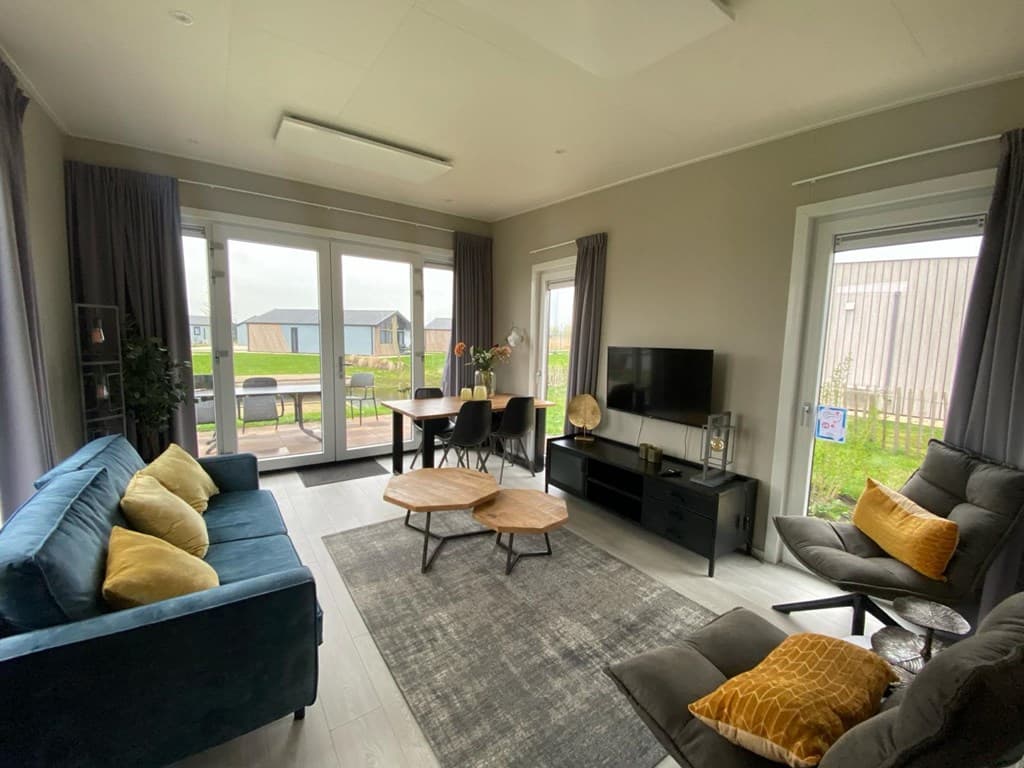 Moderne woonkamer met blauwe bank, eettafel, TV-meubel en grote ramen met uitzicht op buiten.