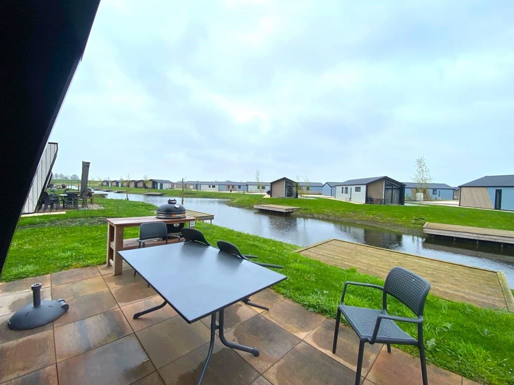 Terras met tafel en stoelen aan water, uitzicht op vakantiewoningen en groene omgeving met gras en paden.