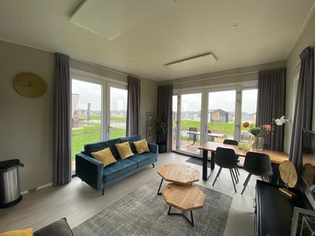 Woonkamer met een blauwe bank, een eettafel en grote ramen met uitzicht op de buitenruimte en andere vakantiewoningen.