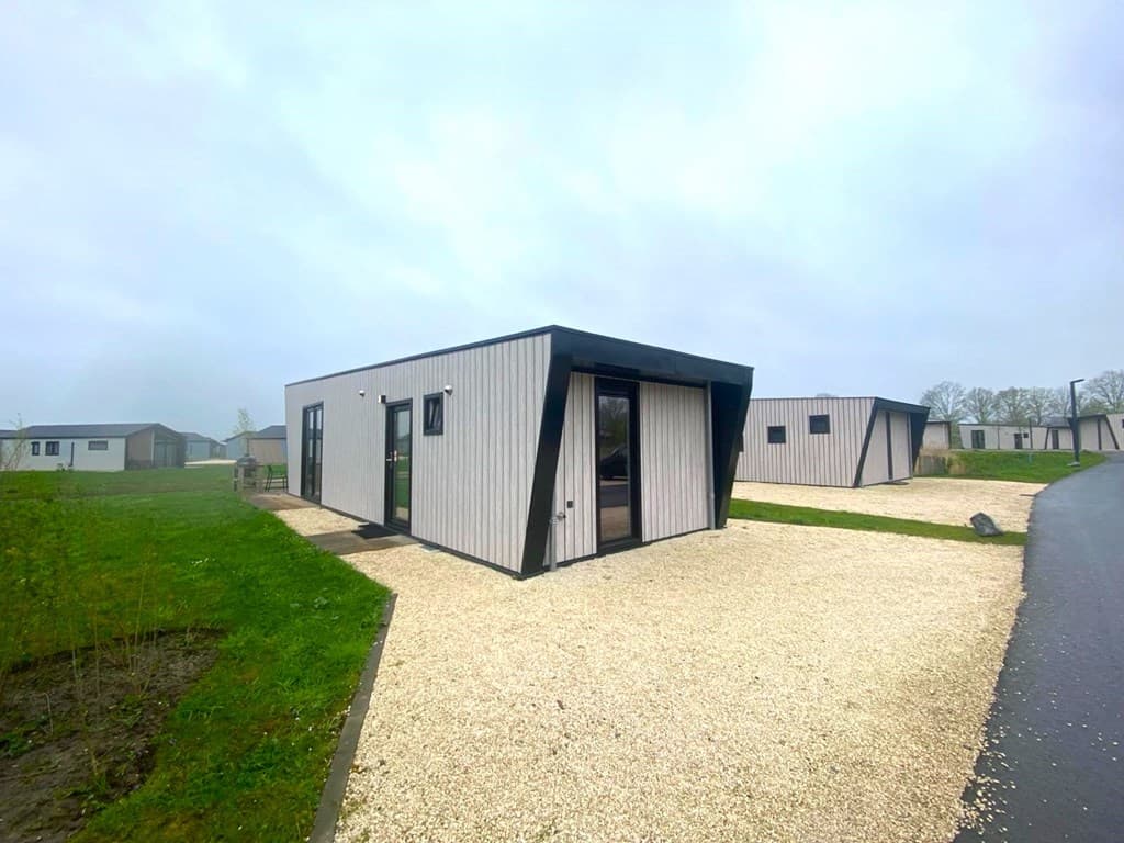 Moderne vakantiewoning met grijze gevel en grote ramen, gelegen op een grindpad met groene omgeving.