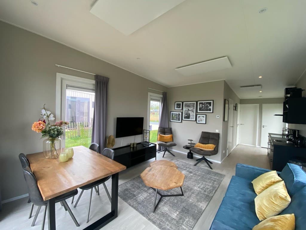 Woonkamer met eettafel, bank en tv, donkere kasten en foto's aan de muur, lichtinval door grote ramen