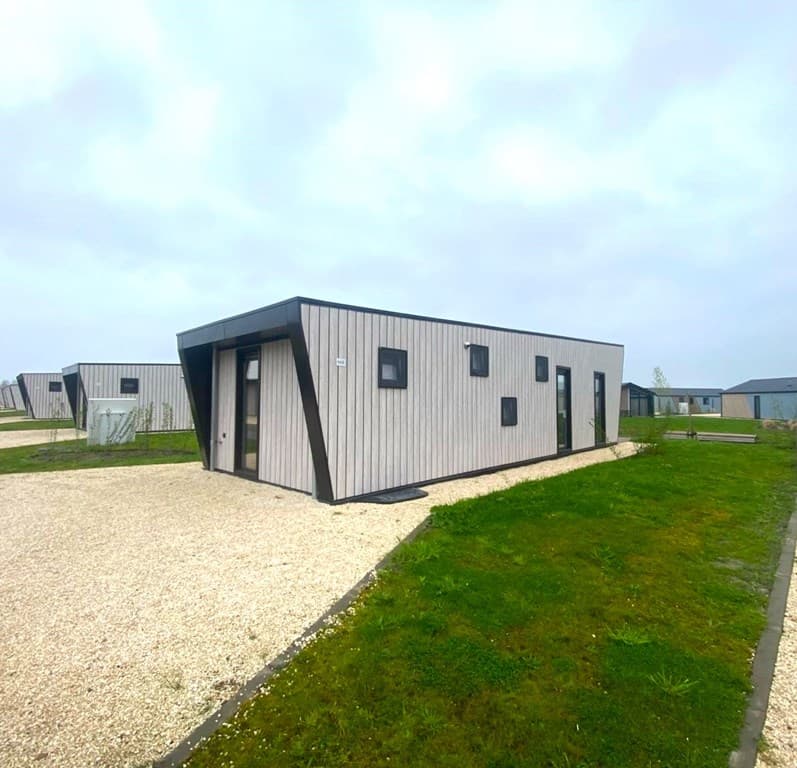 Moderne vakantiewoning met een unieke gevel en een grindpad, omringd door gras en andere woningen.