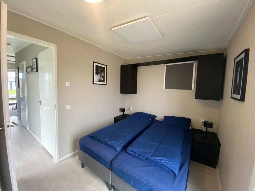 Slaapkamer met twee eenpersoonsbedden, nachtkastjes en zwart-grijze wandplanken. Deur naar gang aan de linkerkant.