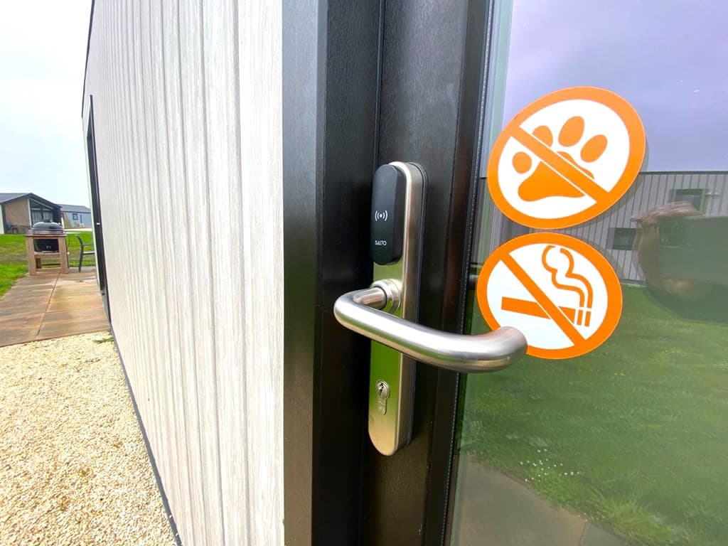 Deurknop van een woning met twee zorgmerken voor huisdieren en roken, zicht op gras en andere woningen op de achtergrond