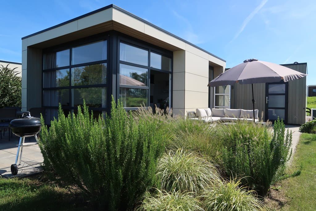 Moderne vakantiewoning met grote ramen, terras en een parasol, omringd door groene planten en een barbecue.