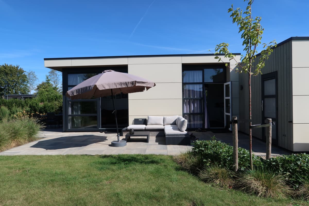 Moderne vakantiewoning met een loungeset en parasol op een grasveld, omringd door groene begroeiing.