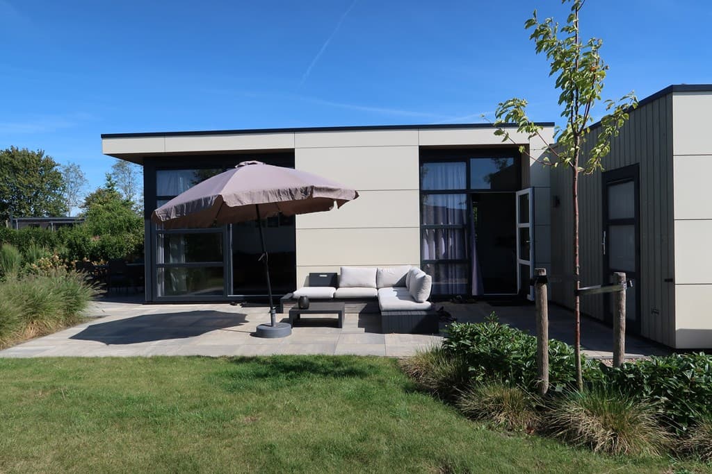 Moderne vakantiewoning met een loungeset en parasol op een grasveld, omringd door groene begroeiing.