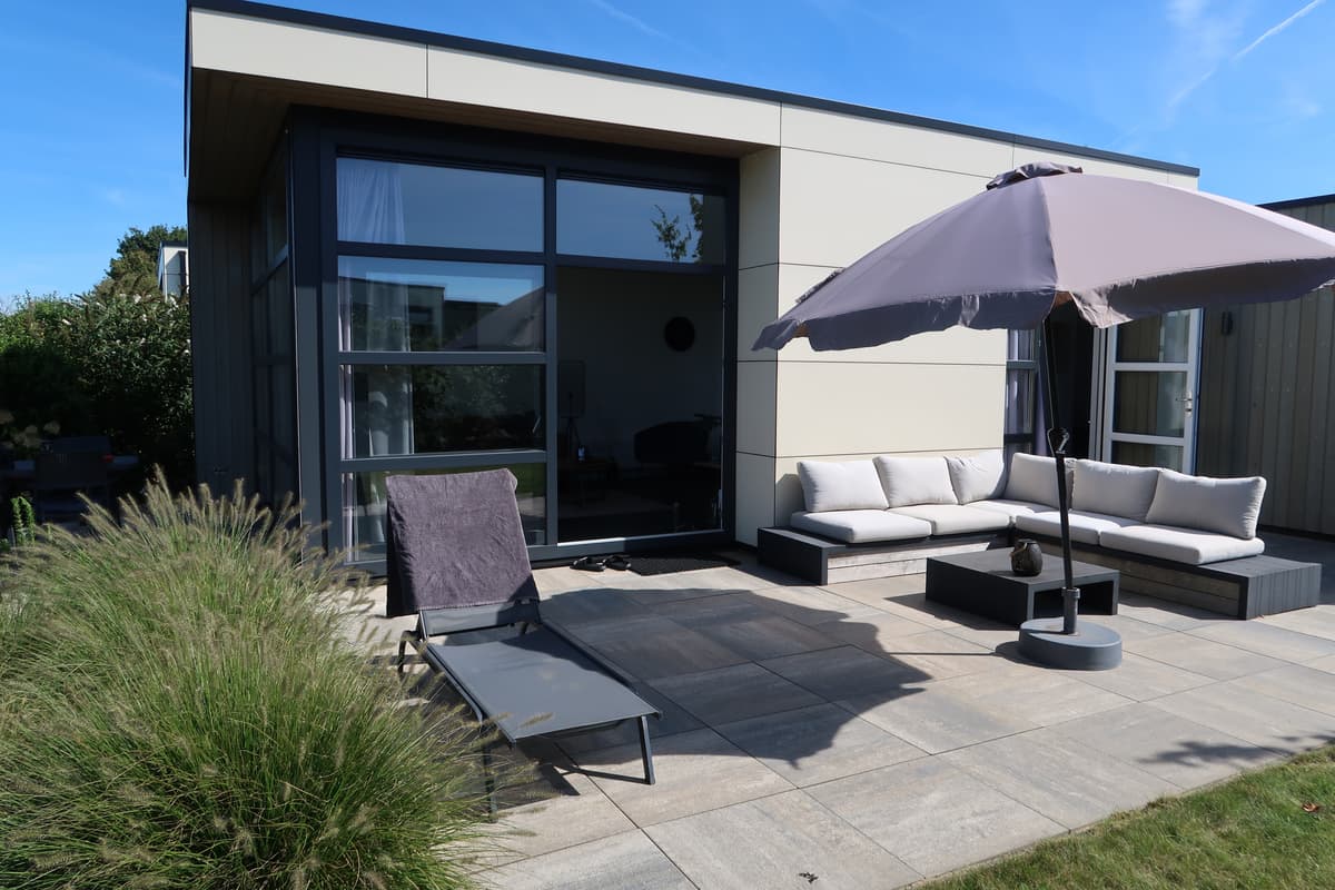 Terras met loungeset en ligstoel, omgeven door groen, onder een parasol met helderblauwe lucht.