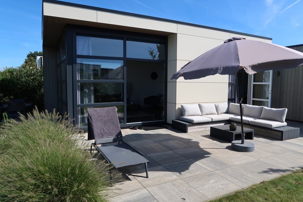 Terras met loungeset en ligstoel, omgeven door groen, onder een parasol met helderblauwe lucht.