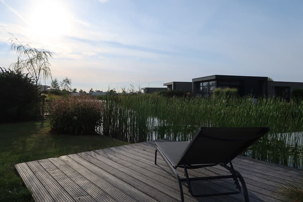 Zicht op een ontspannen ligstoel aan een tuin met riet langs het water en moderne woningen op de achtergrond