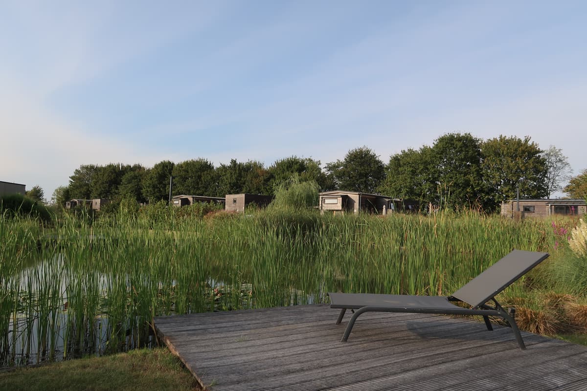 Zicht op vakantiehuisjes vanaf een houten terrace met ligstoel en begroeiing rondom een vijver
