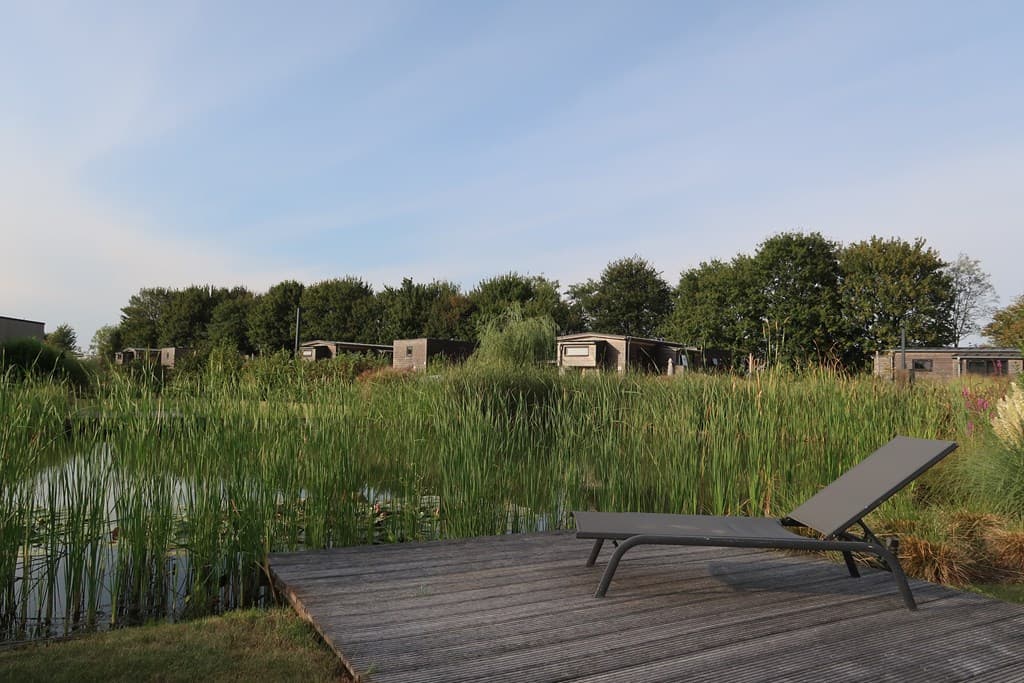Zicht op vakantiehuisjes vanaf een houten terrace met ligstoel en begroeiing rondom een vijver