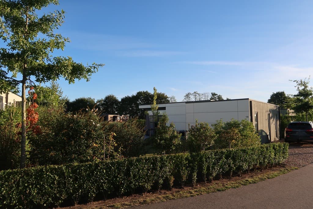 Vakantiewoning omgeven door groen en struiken, met een blauwe lucht op de achtergrond en een brede oprit.