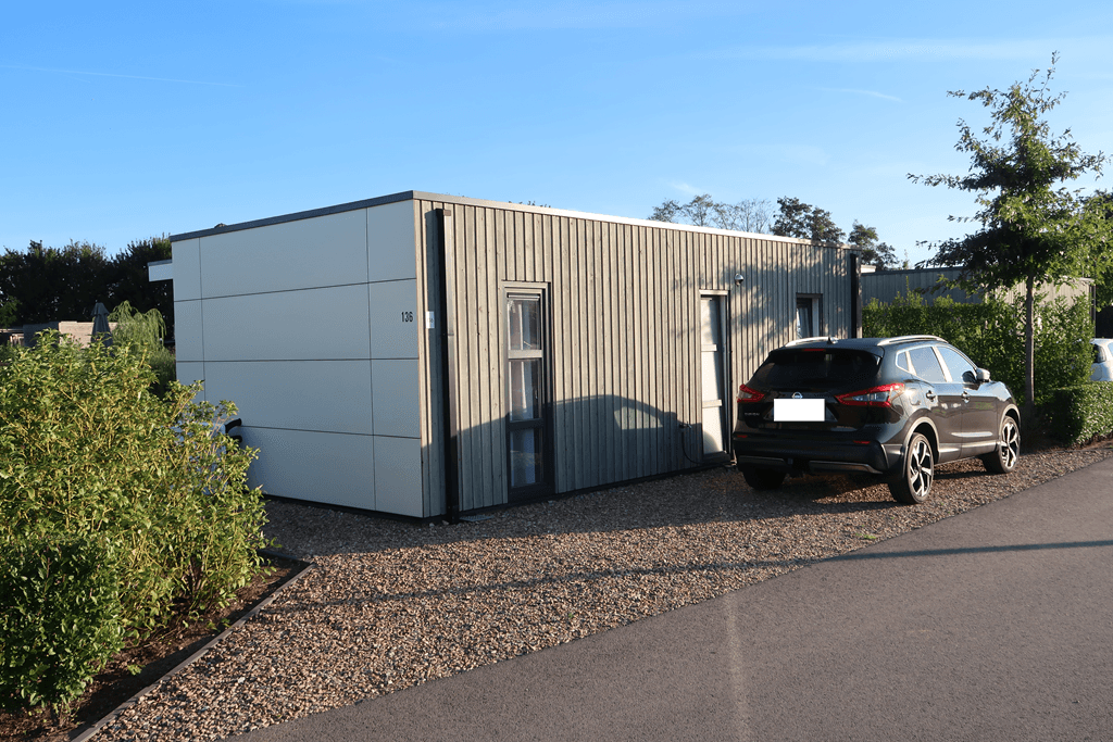 Moderne vakantiewoning met houten gevel en parkeerplaats voor een auto, omringd door groen en gravelpad
