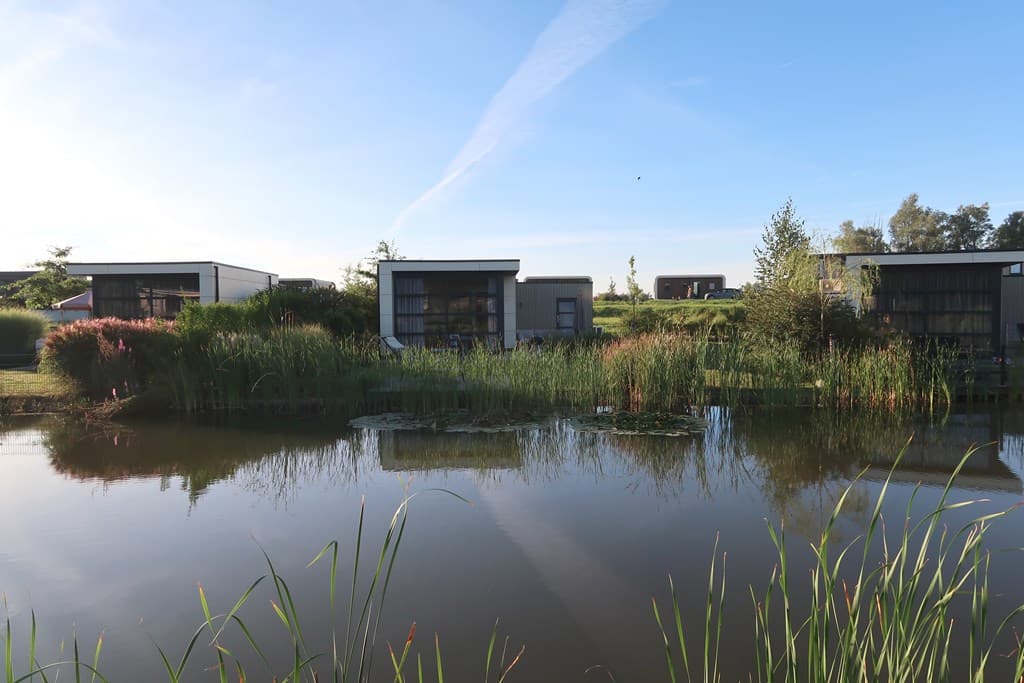 Moderne vakantiewoningen aan een waterpartij omringd door groen en begroeiing onder een blauwe lucht