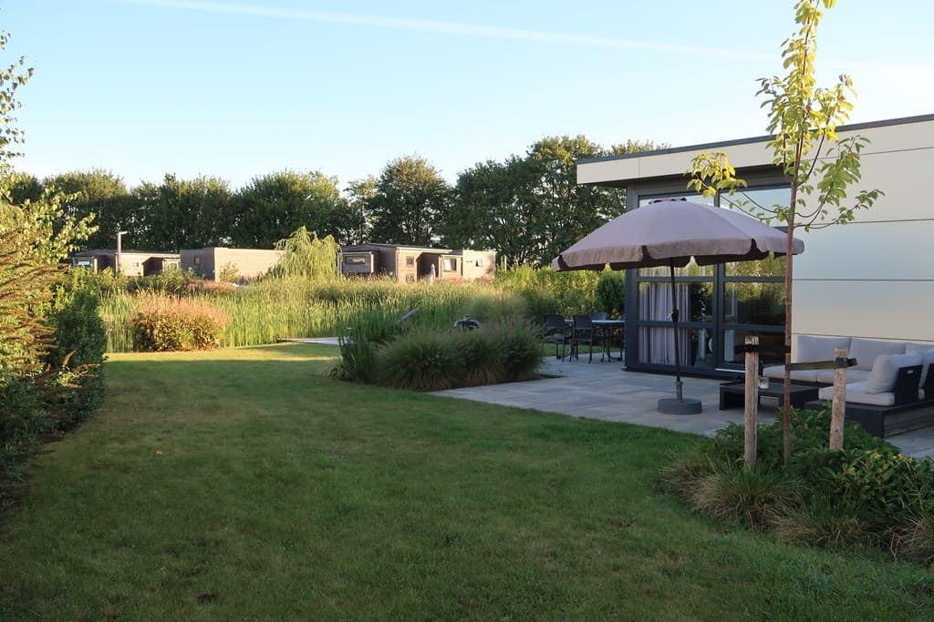 Tuin met groene grasvelden, terrasje met parasol en uitzicht op vakantiehuizen achterin het vakantiepark