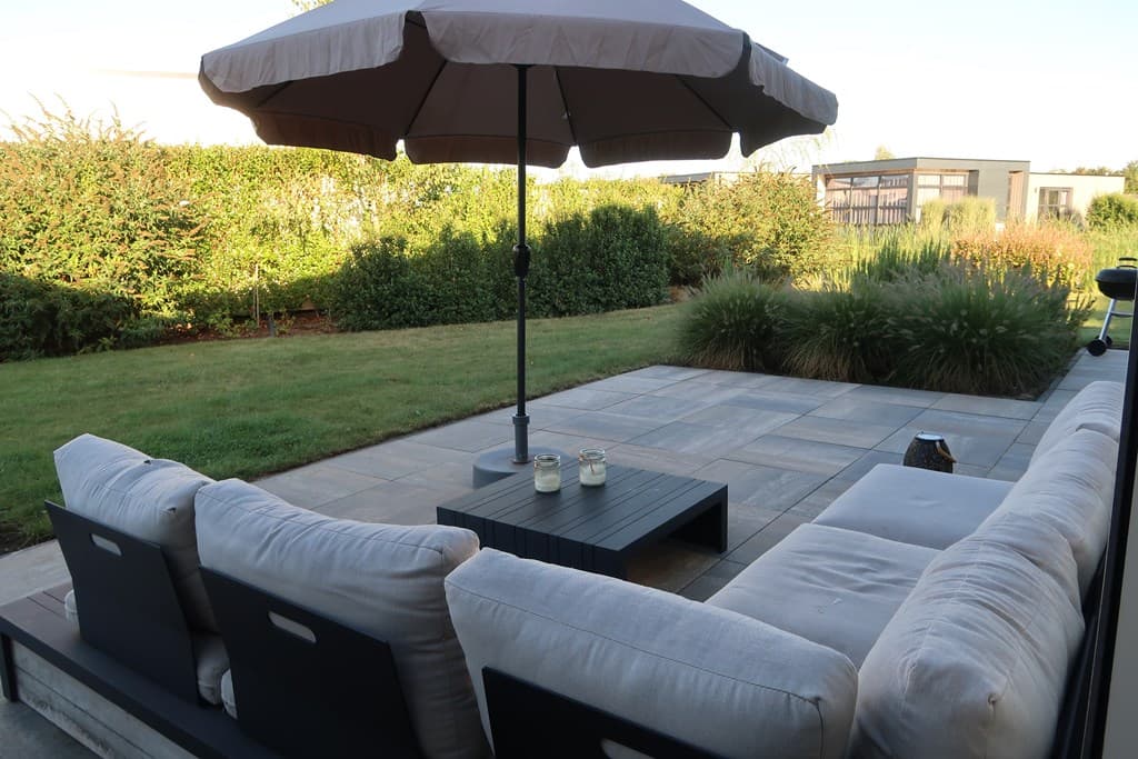 Terras met een zitgedeelte, een parasol en een groene tuin met struiken en gras rondom.