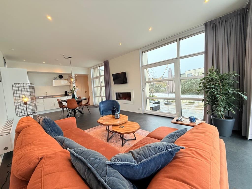Moderne woonkamer met een oranje bank, glazen deuren naar buiten en moderne eethoek in de achtergrond.