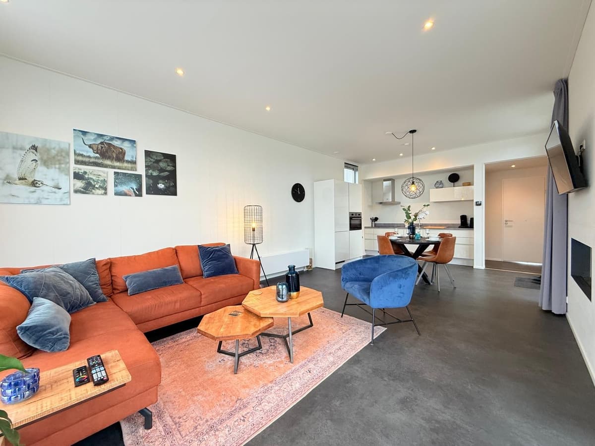 Hedendaagse woonkamer met oranje bank, blauwe stoelen en open keuken, sfeervol ingericht met kunst aan de muur.