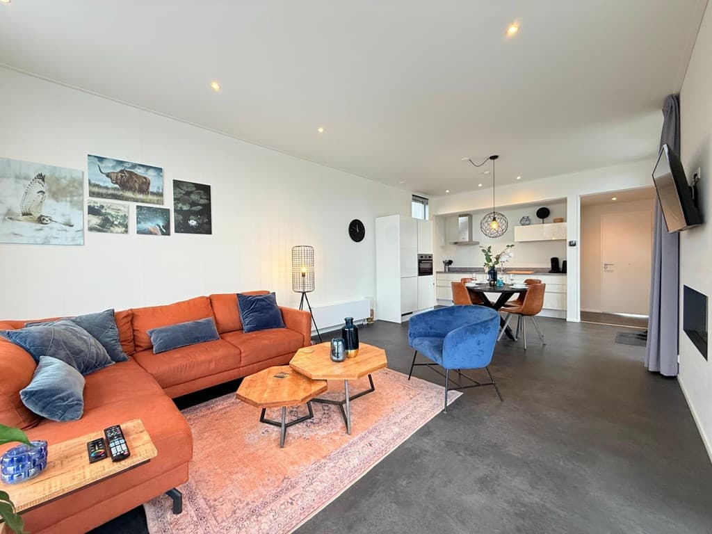 Hedendaagse woonkamer met oranje bank, blauwe stoelen en open keuken, sfeervol ingericht met kunst aan de muur.