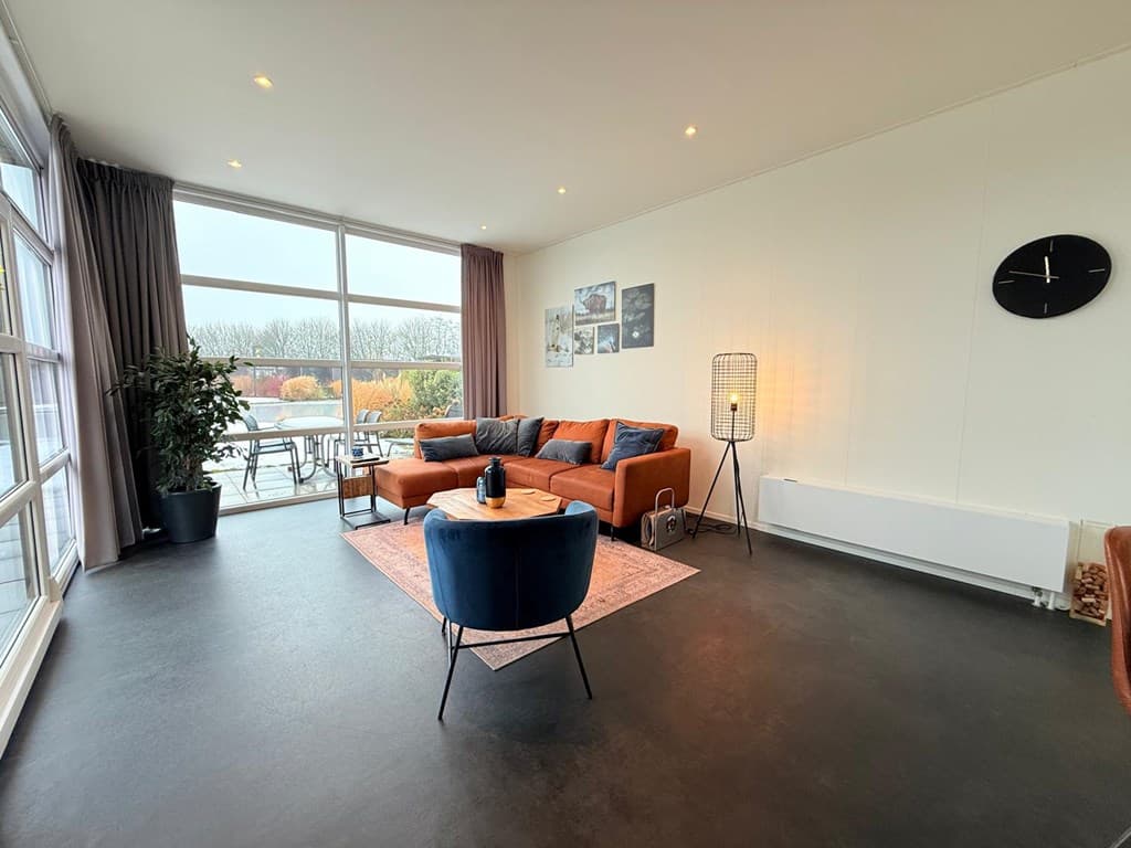 Moderne woonkamer met oranje banken, een tapijt, een houten tafel en grote ramen met uitzicht op het buitengebied.