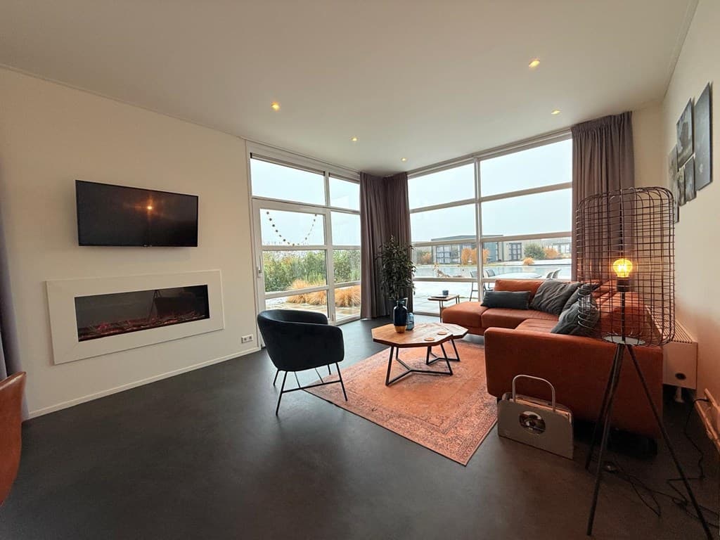 Moderne woonkamer met grote ramen, comfortabele bank, fauteuils, tv en elektrische haard, met uitzicht op buitenruimte.