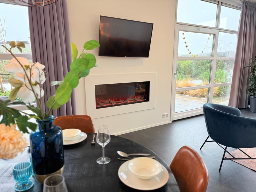 Modern interieur met eettafel, tv boven een elektrische haard en grote ramen met uitzicht op een tuin.