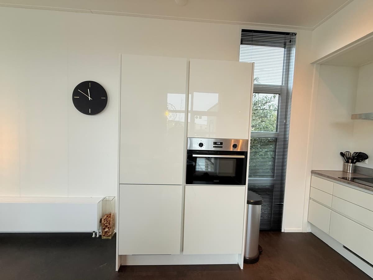 Moderne keuken met witte kasten, oven en koelkast, voorzien van een klok aan de muur en grote ramen voor natuurlijk licht