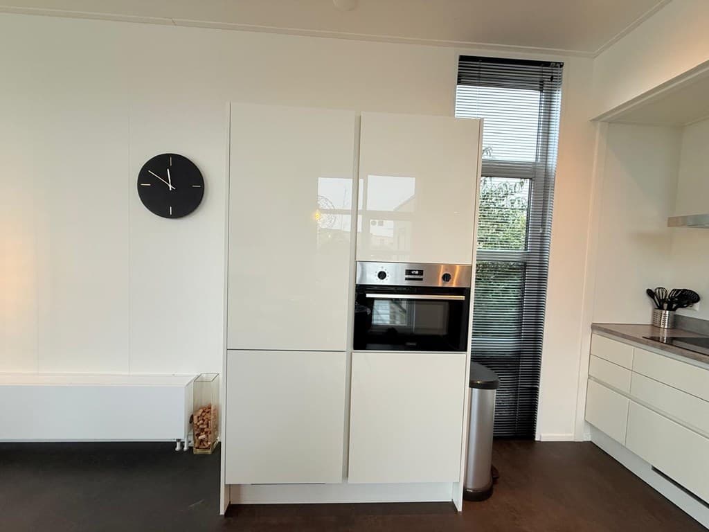Moderne keuken met witte kasten, oven en koelkast, voorzien van een klok aan de muur en grote ramen voor natuurlijk licht