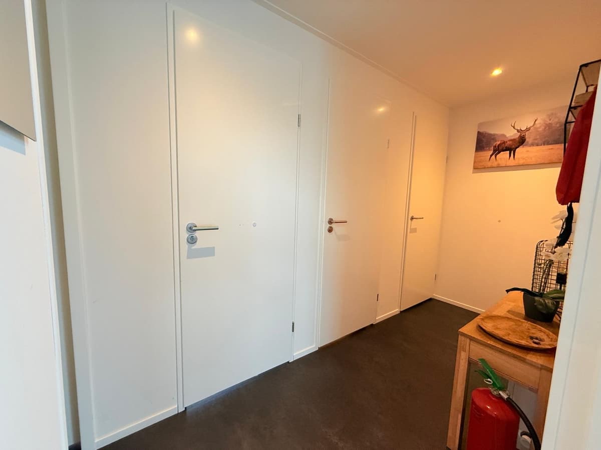 In een lichte hal met witte wanden en drie deuren aan de rechterzijde, een houten tafel met een mand en een schilderij aan de
