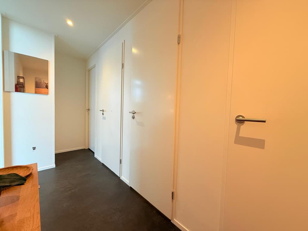 Een gang met witte muren en een donkere vloer, voorzien van deuren aan beide zijden en een spiegel aan de muur.