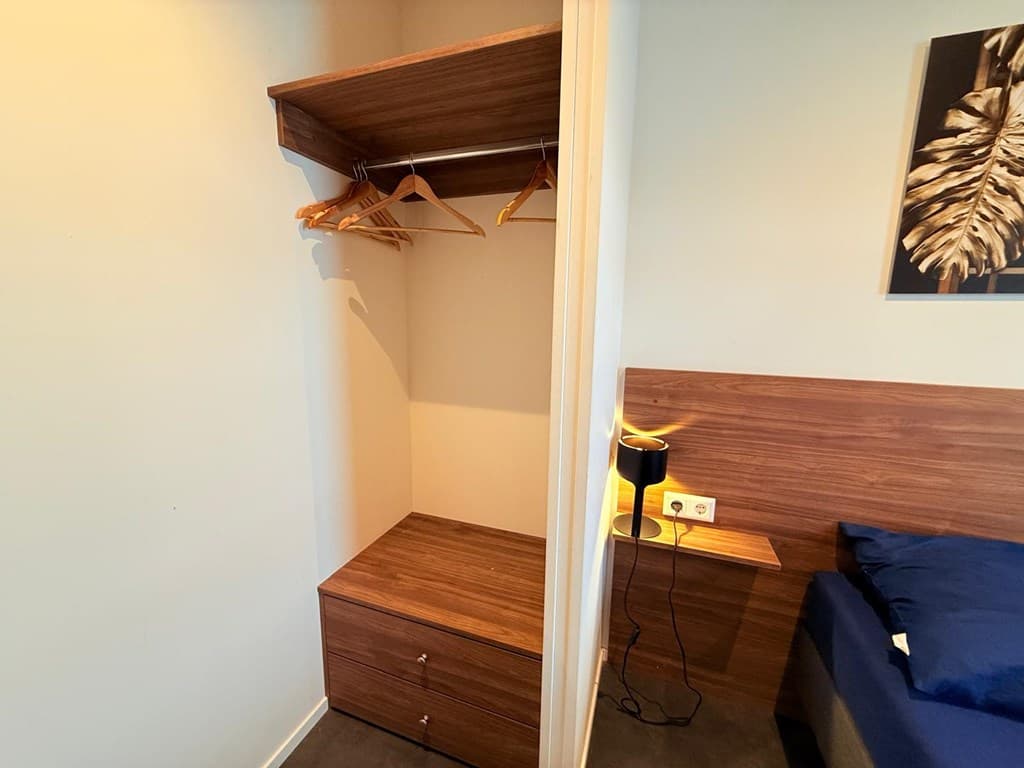 Ingebouwde kast met hangruimte en lades naast een bed en een nachtlampje aan de muur.