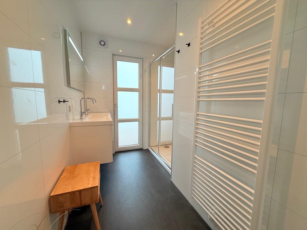 Moderne badkamer met douche, wastafel en spiegel, en een houten bankje bij de deur.
