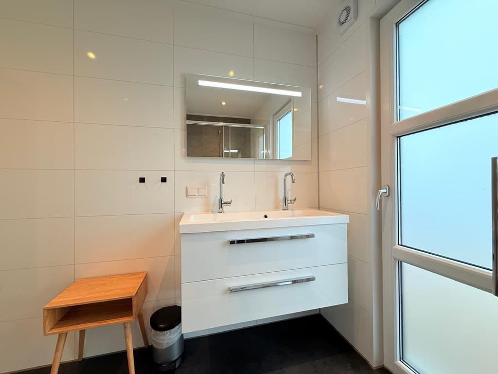 Moderne badkamer met wastafel met dubbele kranen, spiegel, houten krukje en veel daglicht vanuit een raam.
