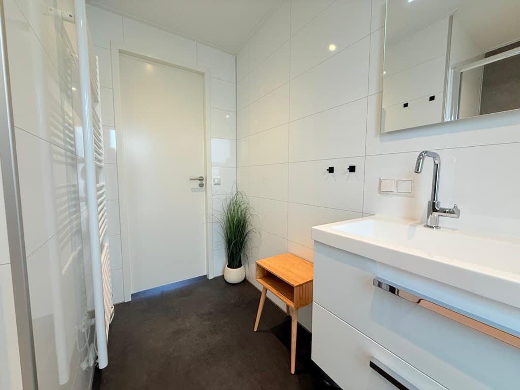 Moderne badkamer met witte wandtegels, houten bankje en wasbak met spiegel, planten in de hoek en lichte inrichting