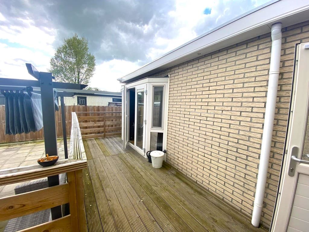Houten terras met een stenen muur, een deur naar binnen en een pergola met zitgelegenheid.