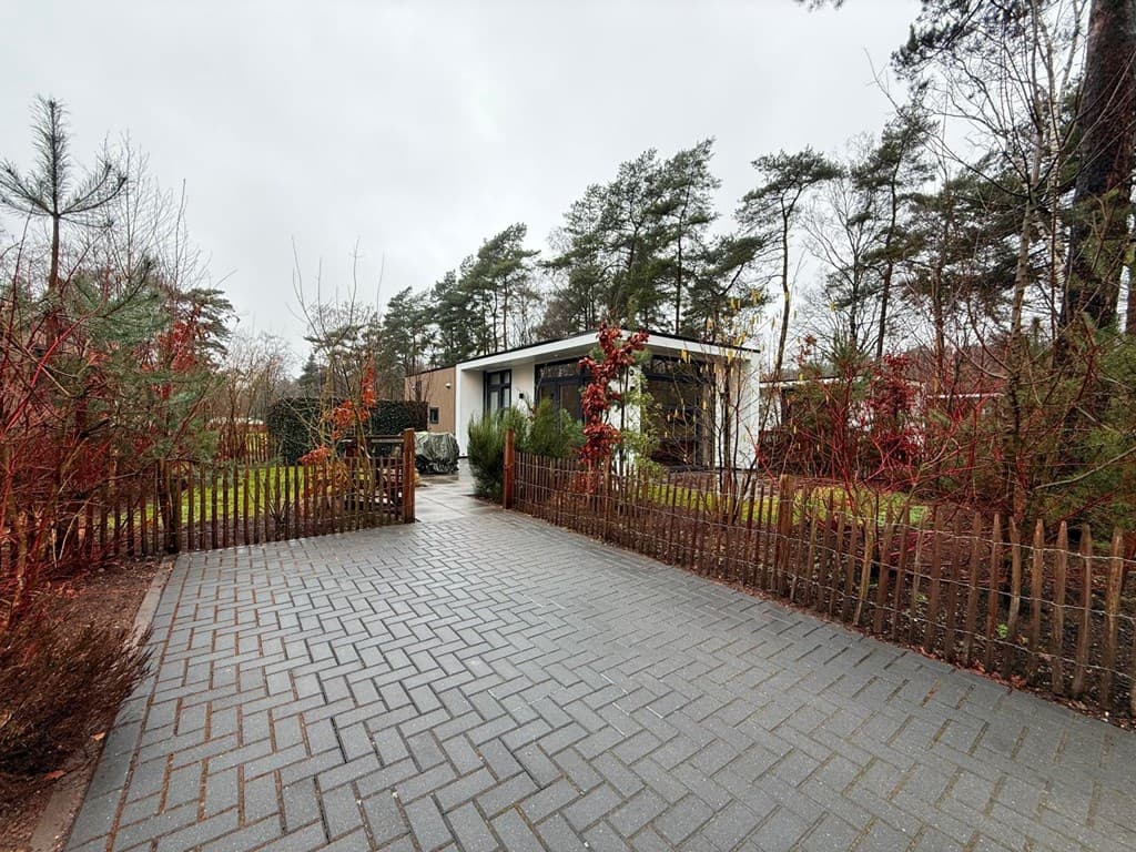Moderne vakantiewoning omringd door bomen en kleurrijke struiken, met een bestrate oprit en houten hekwerk.