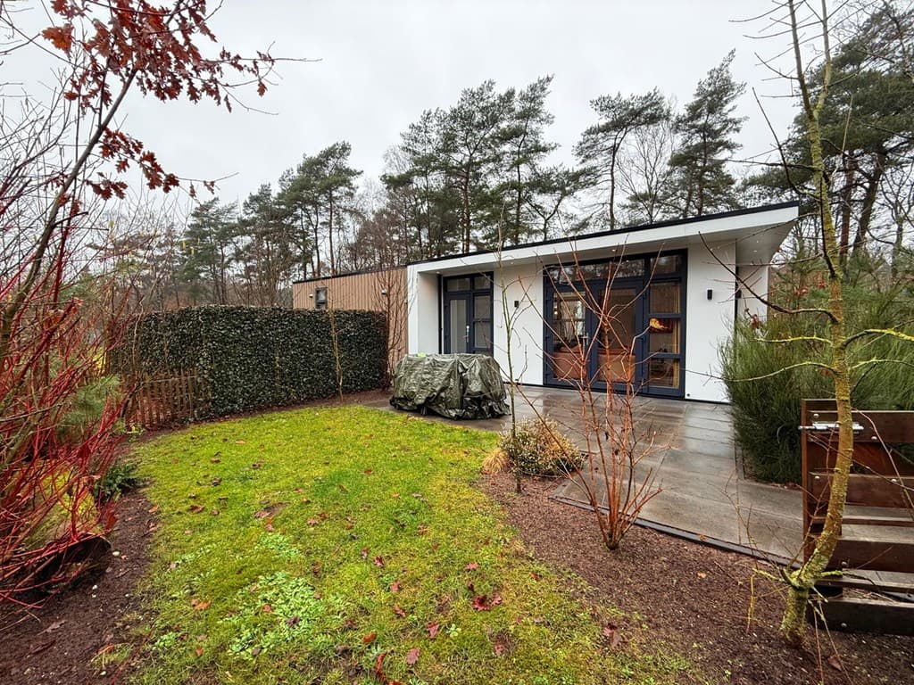 Moderne vakantiewoning met een tuin, omgeven door bomen en struiken, en een terras met een afgedekt meubelstuk.