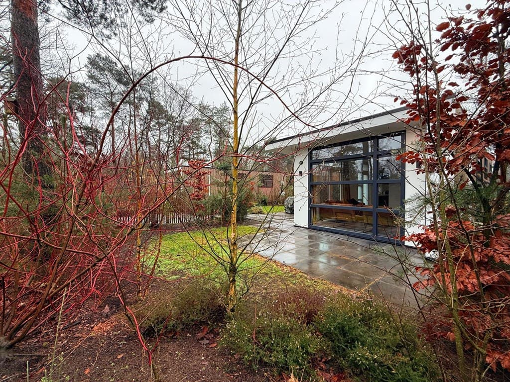 Moderne vakantiewoning omringd door bomen en struiken met een betegeld terras en grote ramen.